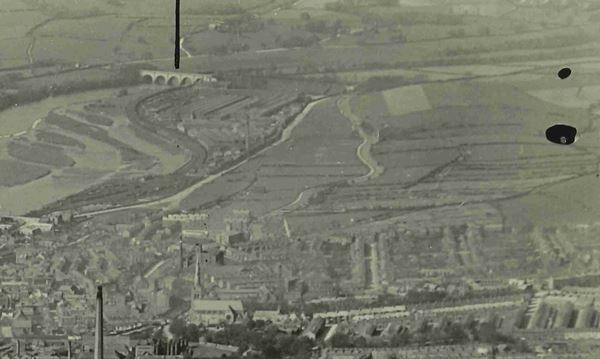 The black and white aerial photograph looks across an housing estate with rows of terraced house in the foreground and then fields beyond. Coming from the right the road can be seen heading into the top of the middle of the picture with a factory next to it. This is where Newton beck can be found. To the left of the factory is the river, curving away and in the top left can be seen the Lune aqueduct. Up the middle of the fields can be seen the canal, curving slightly as it crosses the fields.